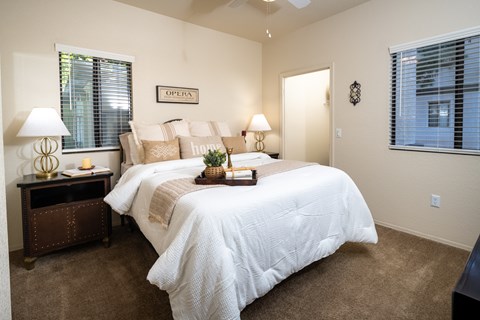 3 Bedroom Apartments in Surprise, AZ - The Village at West Point - A Bedroom With A White Comforter, Neutral Accent Pillows, Matching Nightstands With Lamps, And Two Windows With Blinds.