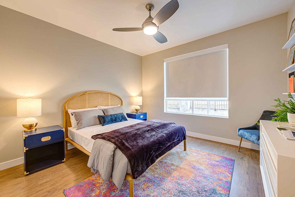 Apartments in Redlands, CA - The Summit - Bedroom With Contemporary Plank Flooring, Ceiling Fan, Window with Roller Shade, Bed, Area Rug, Two Nightstands With Lamps, Chair, and Dresser