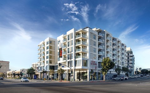 A large white apartment building with a red sign that says "Now Leasing" is under a blue sky.