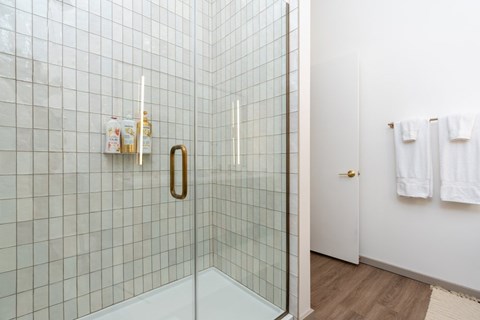 A white tiled shower with a gold handle and a white door with towels hanging on it.