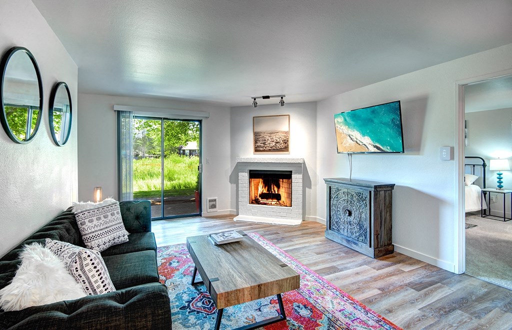 Pet Friendly Apartments in Kirkland WA - Woodlake - Spacious, Furnished Living Room with Wood Flooring, Area Rug, Sofa, Wooden Cofeetable, Wooden Chest, Mounted TV, Fireplace with White Mantel, Wall Art, Mirrors, Sliding Glass Door, and View of Bedroom