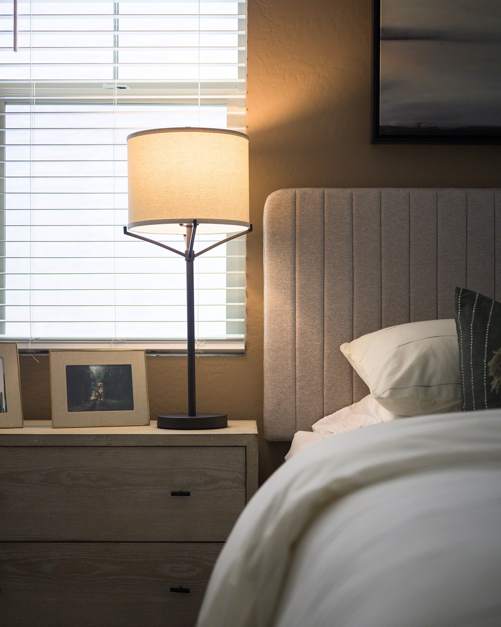 A bedroom with a bed, nightstand with a lamp and picture frame, and a window with blinds.