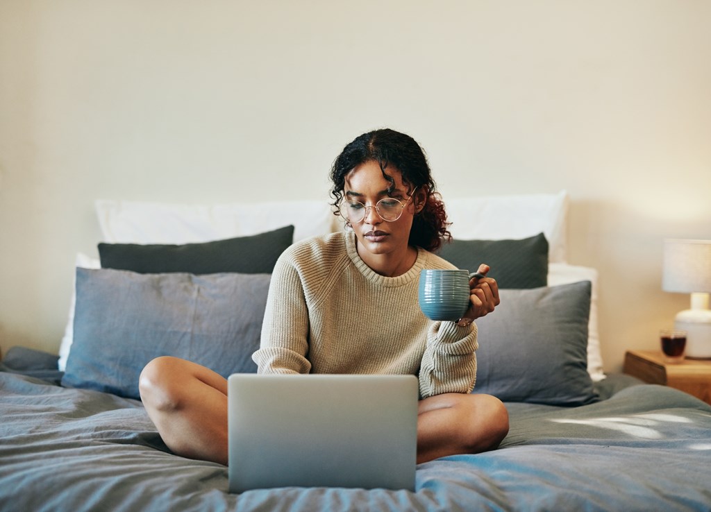 A person is sitting on a bed with a laptop and holding a mug.