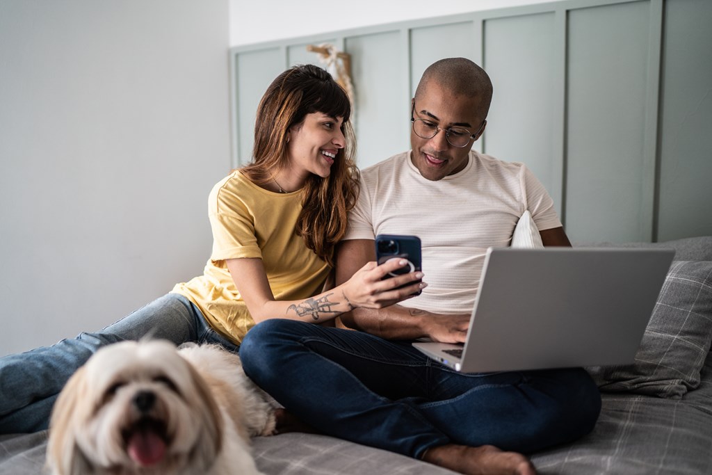 A man and woman sitting on a bed with a laptop and a dog, both looking at a phone.
