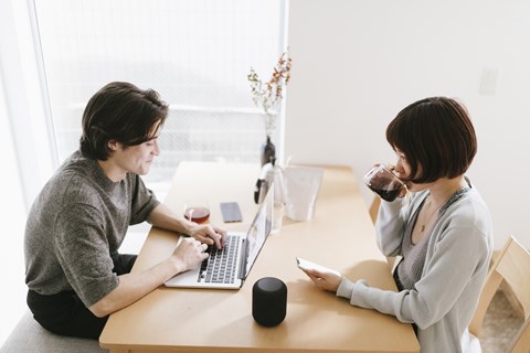 A man and a woman are sitting at a table with a laptop, a smart speaker, and a smartphone.