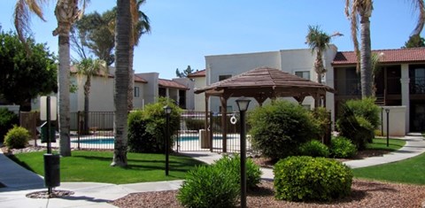 A gazebo is surrounded by bushes and palm trees in front of a building.