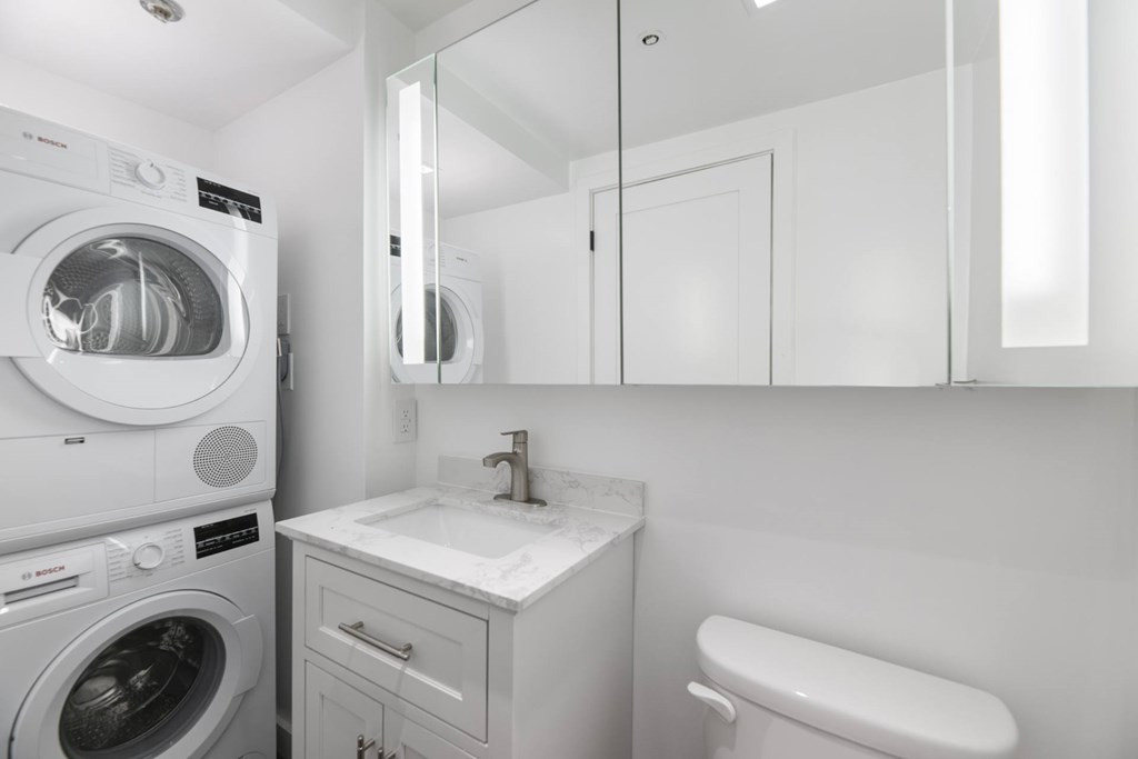 A white washing machine is in a small laundry room with a sink and mirror.