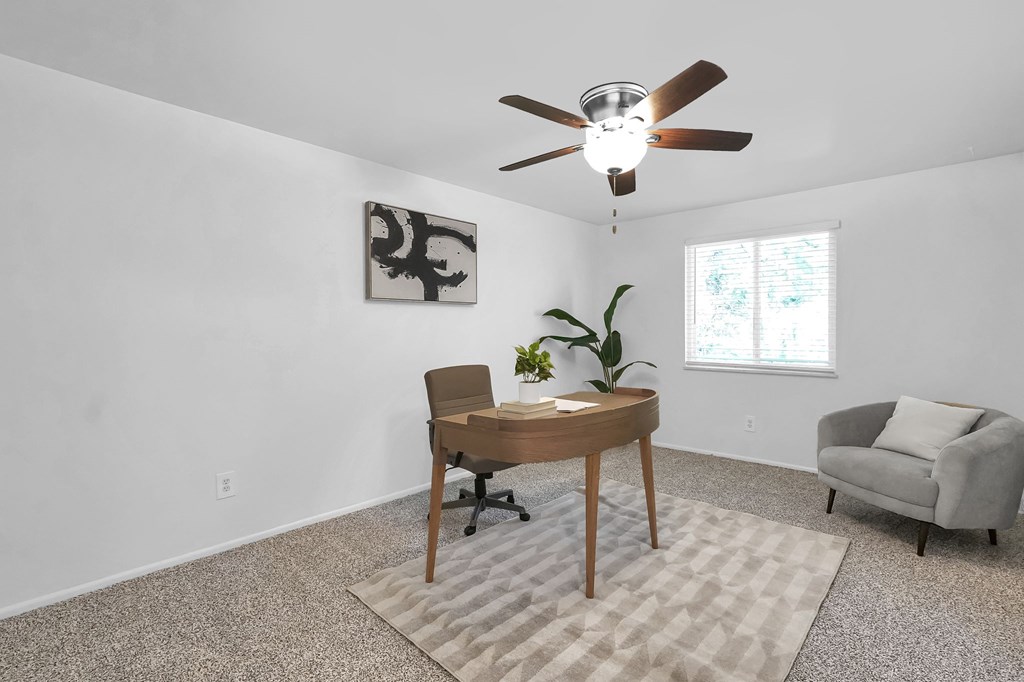 A room with a ceiling fan, a table with a chair, and a painting on the wall.