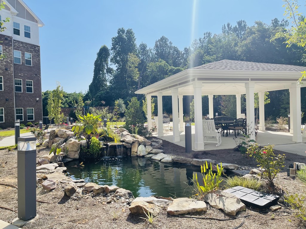 A gazebo is situated in a garden with a pond and a building in the background.