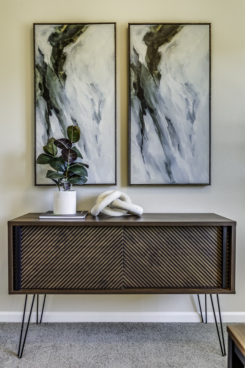 A black and white abstract painting hangs on a wall above a black cabinet with a plant on top.