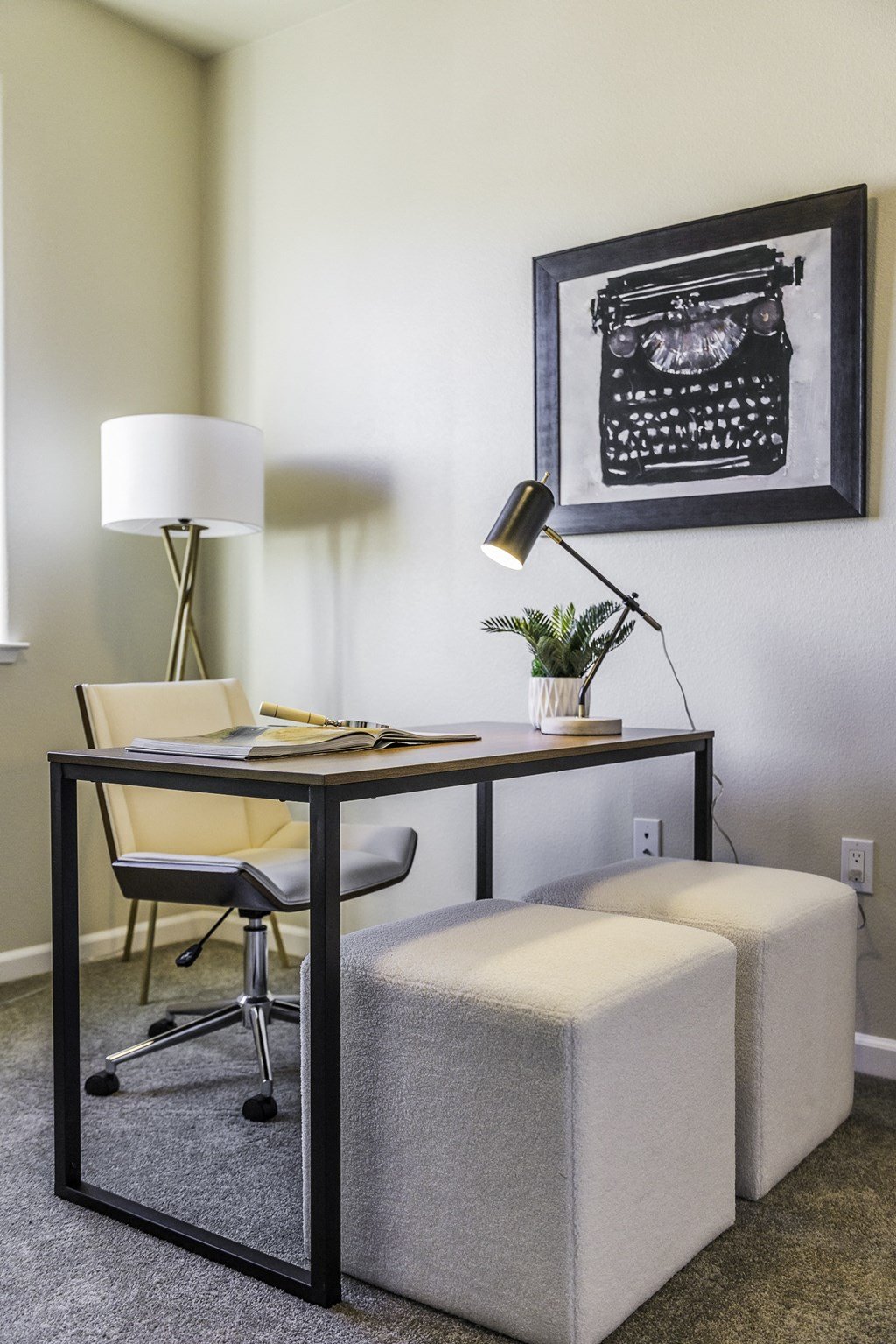 A black and white picture of a typewriter is hanging on a wall above a desk with a chair and a lamp.