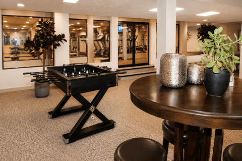 A foosball table is in the middle of a carpeted room with a round table and two potted plants.