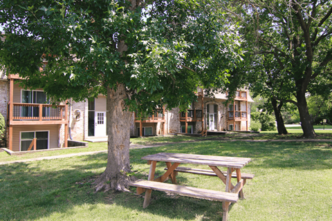 A wooden picnic table is in the foreground of a grassy area with a tree and apartment buildings in the background.
