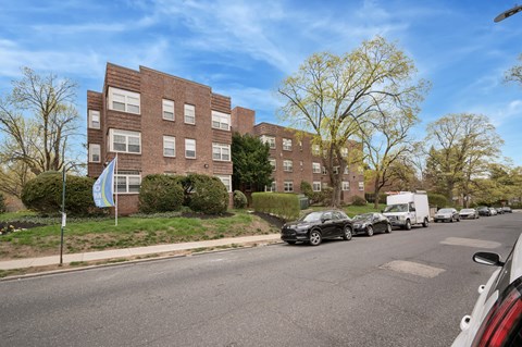 A street view of a residential area with cars parked on the side of the road and apartment buildings in the background.