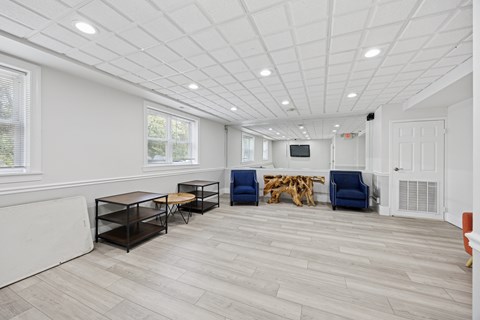 A spacious room with a white ceiling and wooden flooring, furnished with a TV, a couple of chairs, and a table.