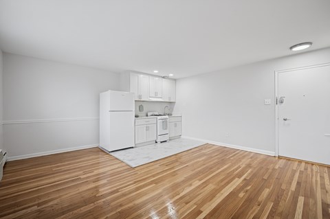A kitchen with white cabinets and a white fridge is in a room with wooden floors and white walls.