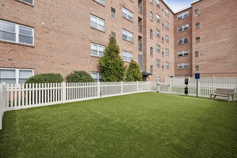 A white picket fence surrounds a green lawn in front of a brick building.