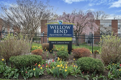 A sign for Willow Bend Apartment Homes is in front of a fence and flowers.