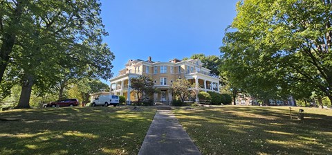 A large house is surrounded by trees and has a driveway leading to the front door.