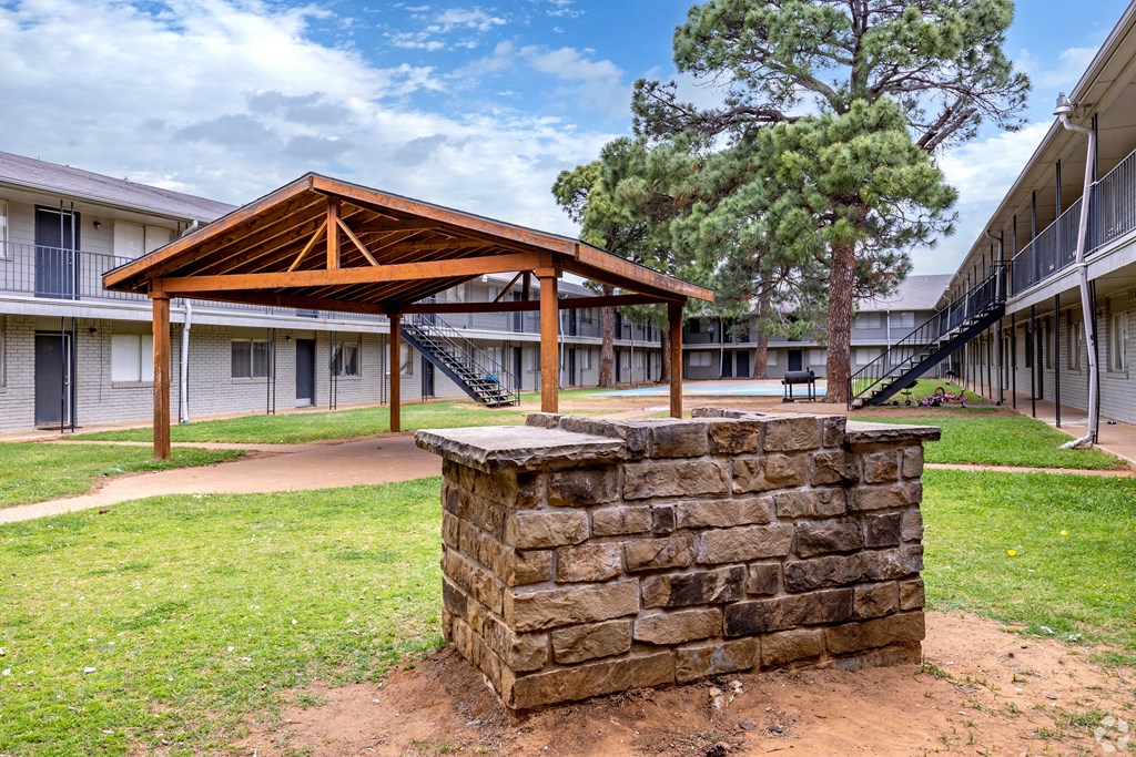 A wooden pavilion is in the middle of a grassy area with a stone pillar in the foreground.