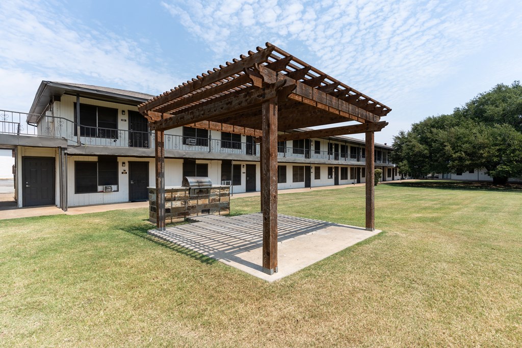 A wooden pergola is in the middle of a grassy area in front of a building.