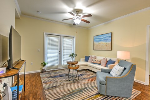 A living room with a grey sofa, a wooden table, a television, a ceiling fan, and a painting of a beach scene on the wall at Cabana Club - Galleria Club Apartments, Jacksonville, 32256
