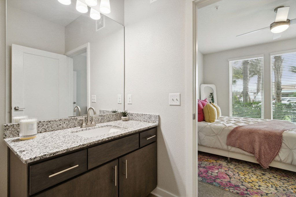 A bathroom with a sink, mirror, and a bedroom with a bed and windowat Pinnacle Apartments, Jacksonville, FL