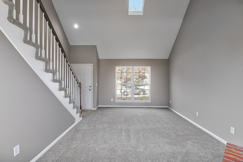 A spacious room with a staircase on the left and a window with a view of a parking lot and trees outside.