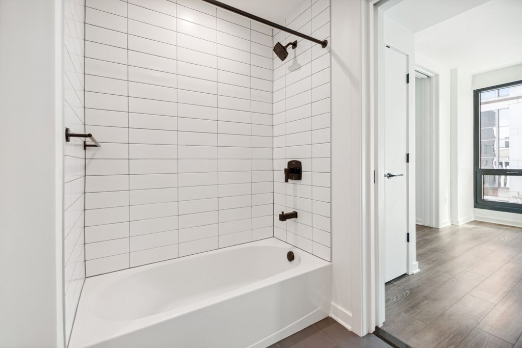 A white tiled bathroom with a white bathtub and a window.
