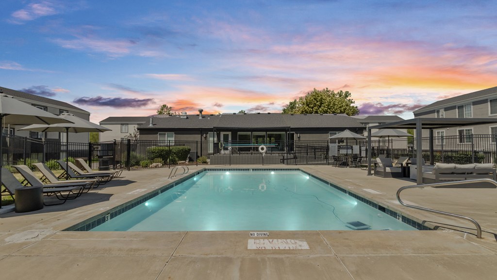 Proximity Apartment Homes pool with chairs and umbrellas and a building with a sunset behind it