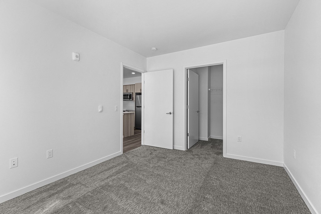 a bedroom with white walls and carpet and a door to a kitchen