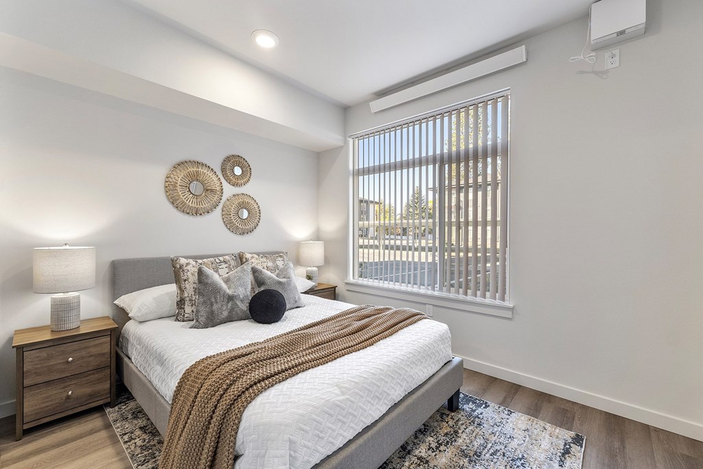 A bedroom with a large bed, a window with blinds, and a nightstand with a lamp.