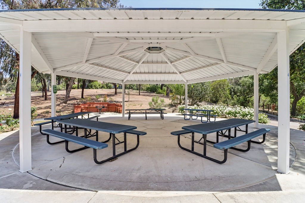 A white pavilion with blue picnic tables is surrounded by trees.