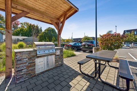 A picnic area with a grill and a table under a wooden roof.