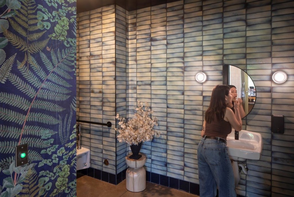 Arts Plaza Gresham Oregon Apartments A woman is brushing her teeth in a bathroom with a wallpapered shower area.