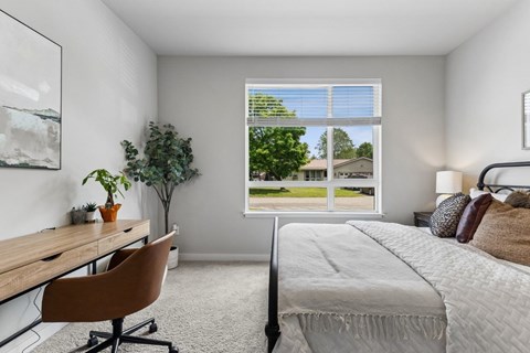 A bedroom with a bed, desk, chair, and a window overlooking a green outdoor area.