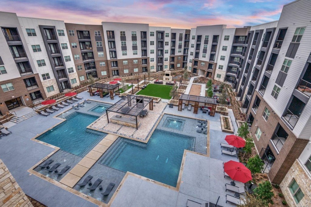 A large swimming pool surrounded by red umbrellas and lounge chairs in a courtyard surrounded by apartment buildings.