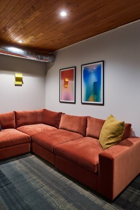 A red couch with a yellow pillow is in the center of a room with two framed pictures on the wall.