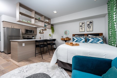 A modern bedroom with a large bed and a kitchen area in the background.