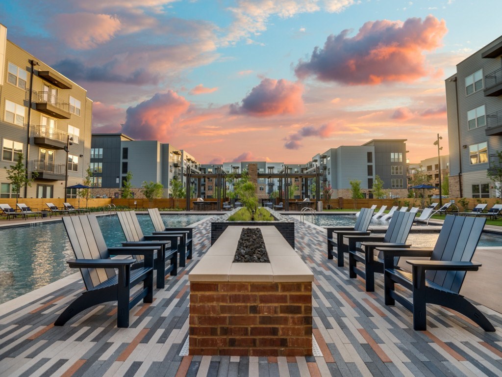 A sunset view of a pool area with benches and a brick fireplace.