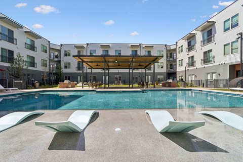 A swimming pool with sun loungers in front of apartment buildings.