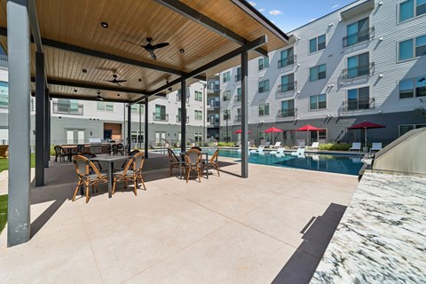 A patio with a table and chairs is surrounded by a pool and buildings.