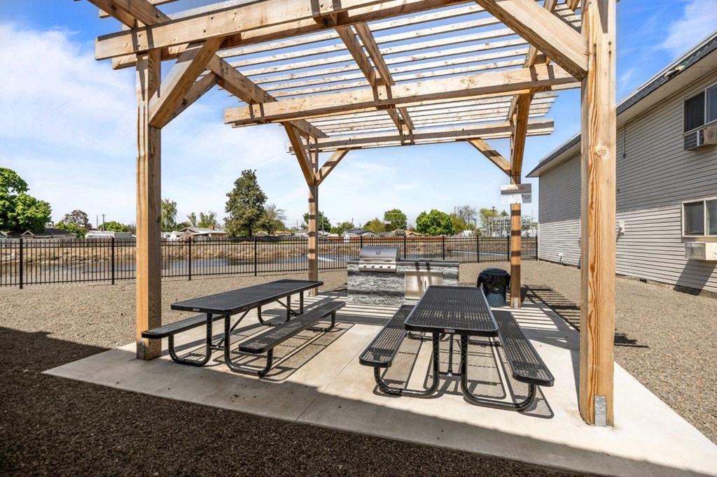 A wooden pergola with picnic tables is set up in a gravel area.