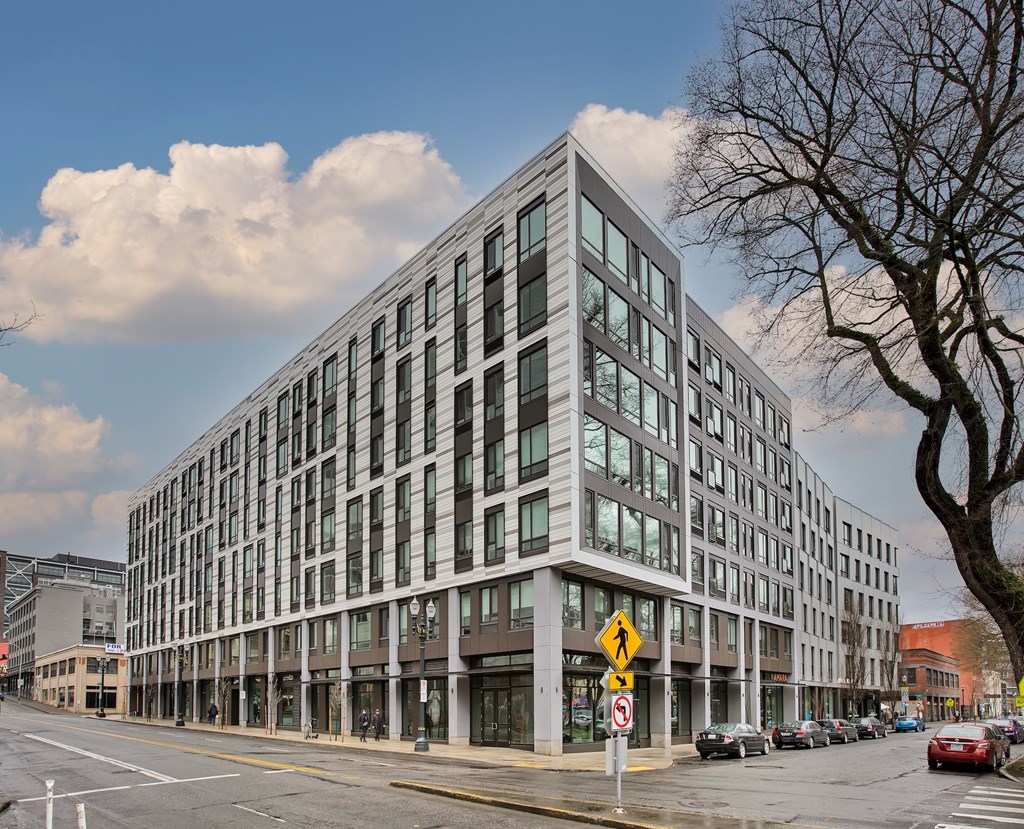 A modern building with a glass facade is situated on a street corner with cars parked along the curb.