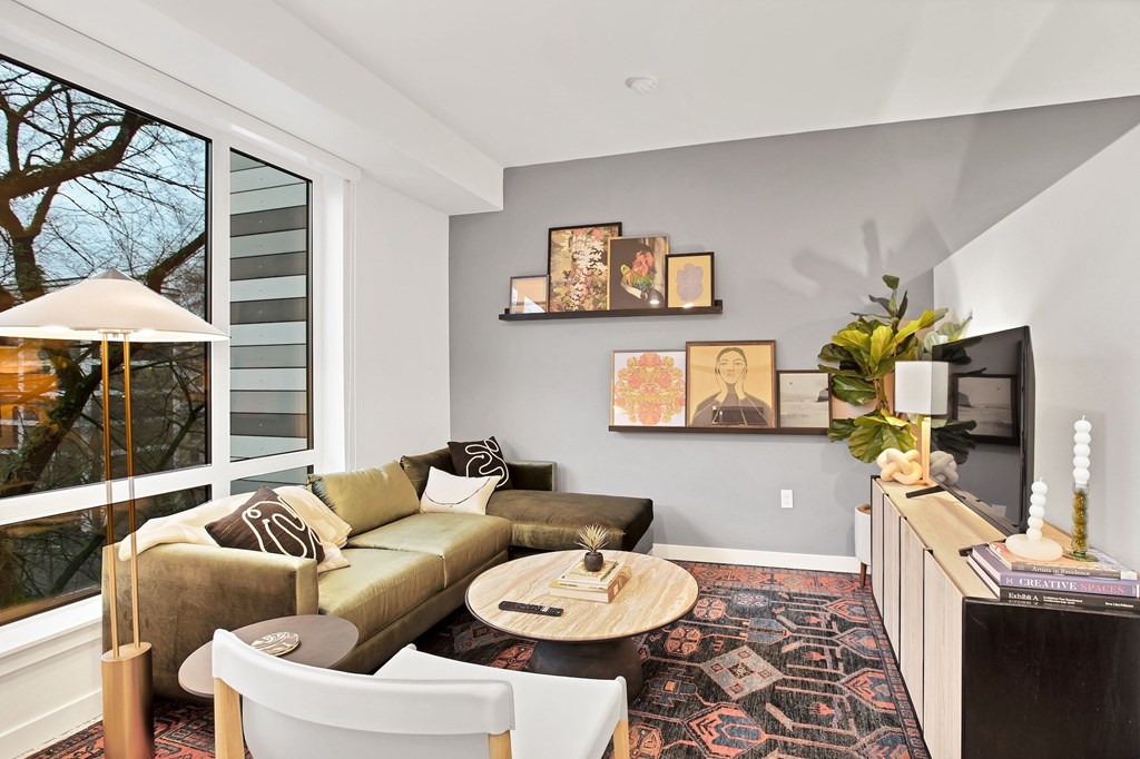 A living room with a couch, a chair, a coffee table, a television, and a large window with a view of a tree.