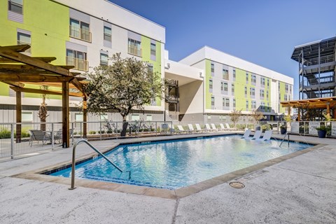 A swimming pool is surrounded by a concrete patio and a building with a yellow and white exterior.