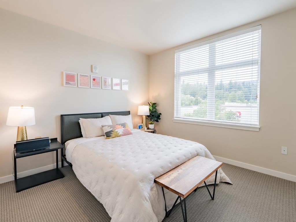 A bedroom with a large bed, a nightstand with a lamp, and a window with blinds.