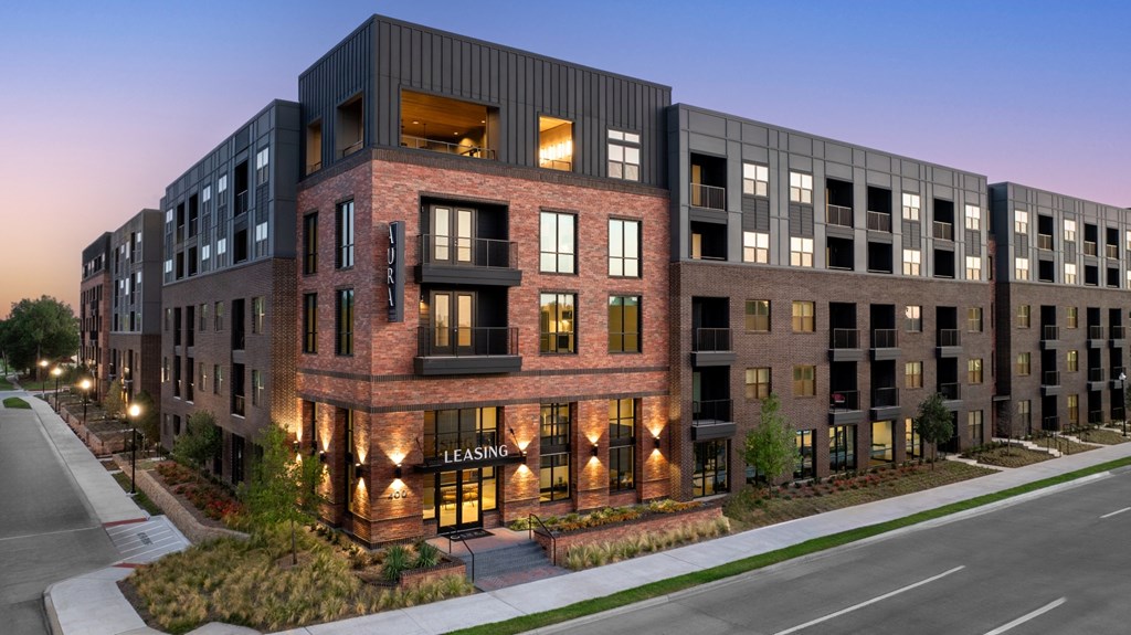 A modern apartment building with a leasing sign is lit up at dusk.