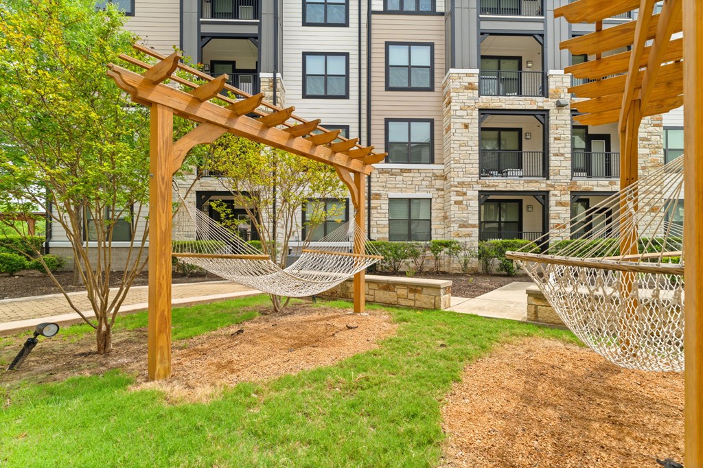 A wooden pergola with a white netting is in the foreground of a well-maintained garden in front of a multi-story apartment building.