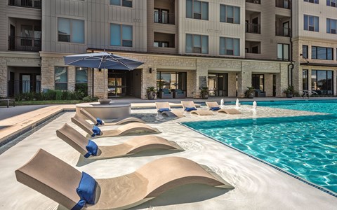 A large swimming pool with lounge chairs and umbrellas in front of a multi-story apartment building.